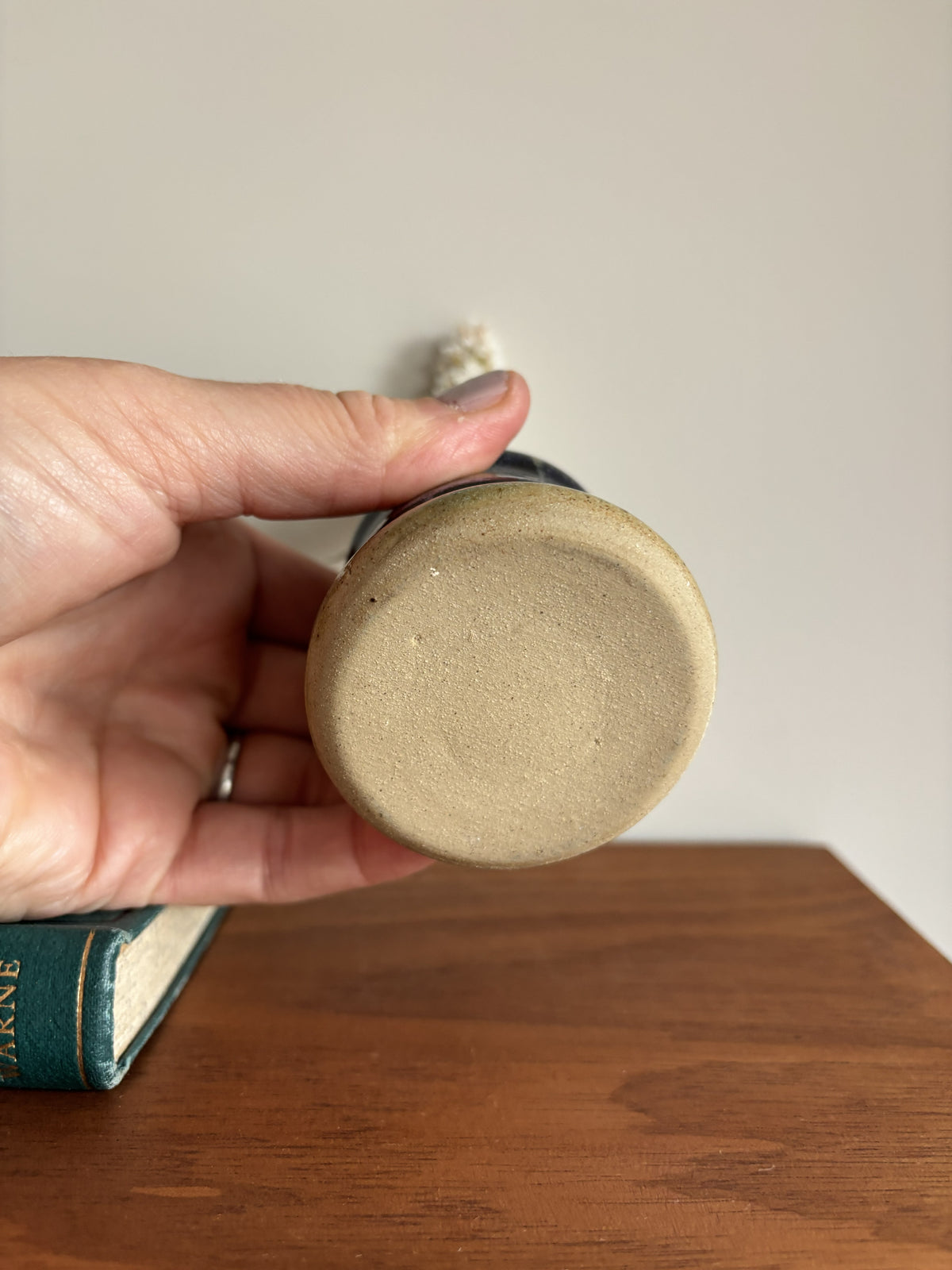 A hand holding the circular base of a ceramic mug or small vessel over a wooden table, with a plain wall behind and part of a book visible at the lower left.