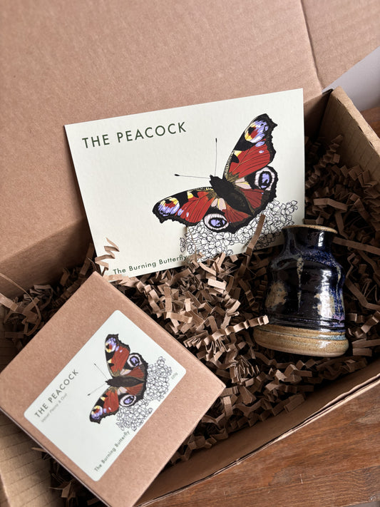 A gift box from The Burning Butterfly containing The Peacock candle in a kraft gift box, a small purple bud vase and The Peacock butterfly A6 postcard. 