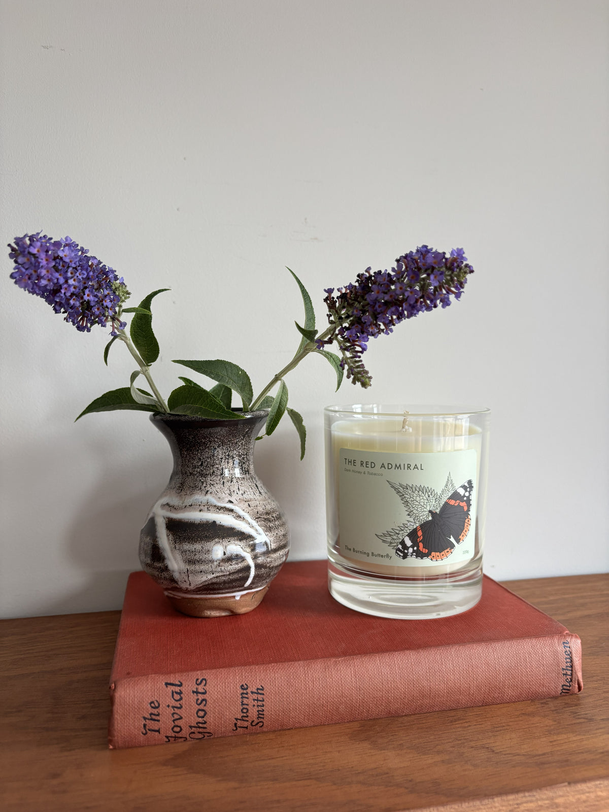 A clear candle with the label The Red Admiral sitting next to a brown and white glazed bud vase with flowers on a red vintage book. 