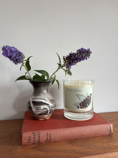 A clear candle with the label The Red Admiral sitting next to a brown and white glazed bud vase with flowers on a red vintage book. 