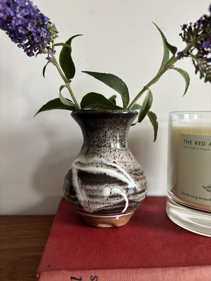 A brown and white bud vase containing purple flowers sitting on top of a red vintage book. 