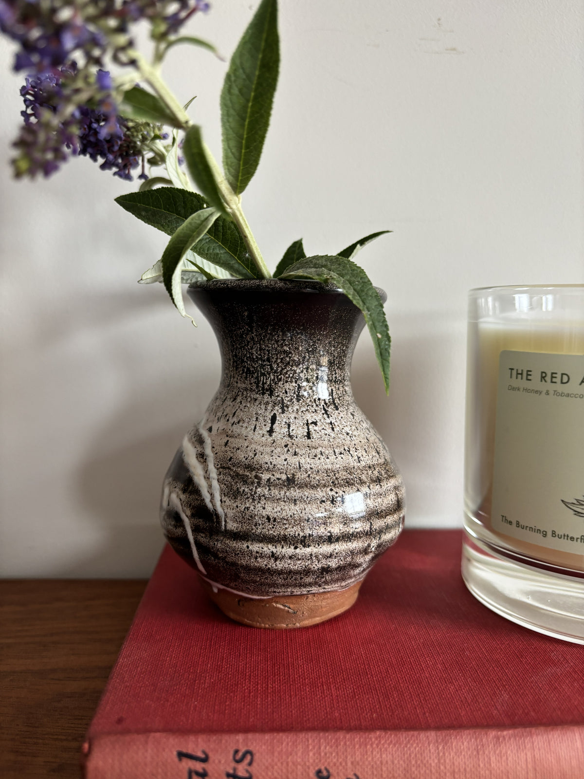 A small handmade ceramic vase with a dark brown and beige speckled glaze, holding green leaves and purple flowers, sitting on a red book next to a candle jar.