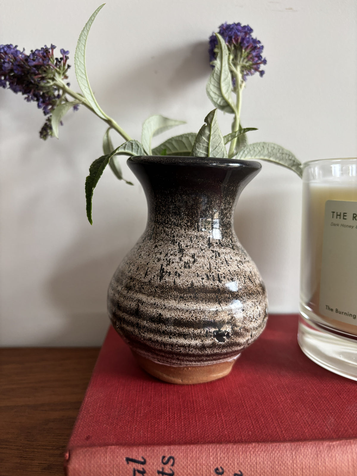 A small handmade ceramic bud vase with a brown and beige speckled glaze, holding green leaves and purple flowers, styled on a red book next to a candle.