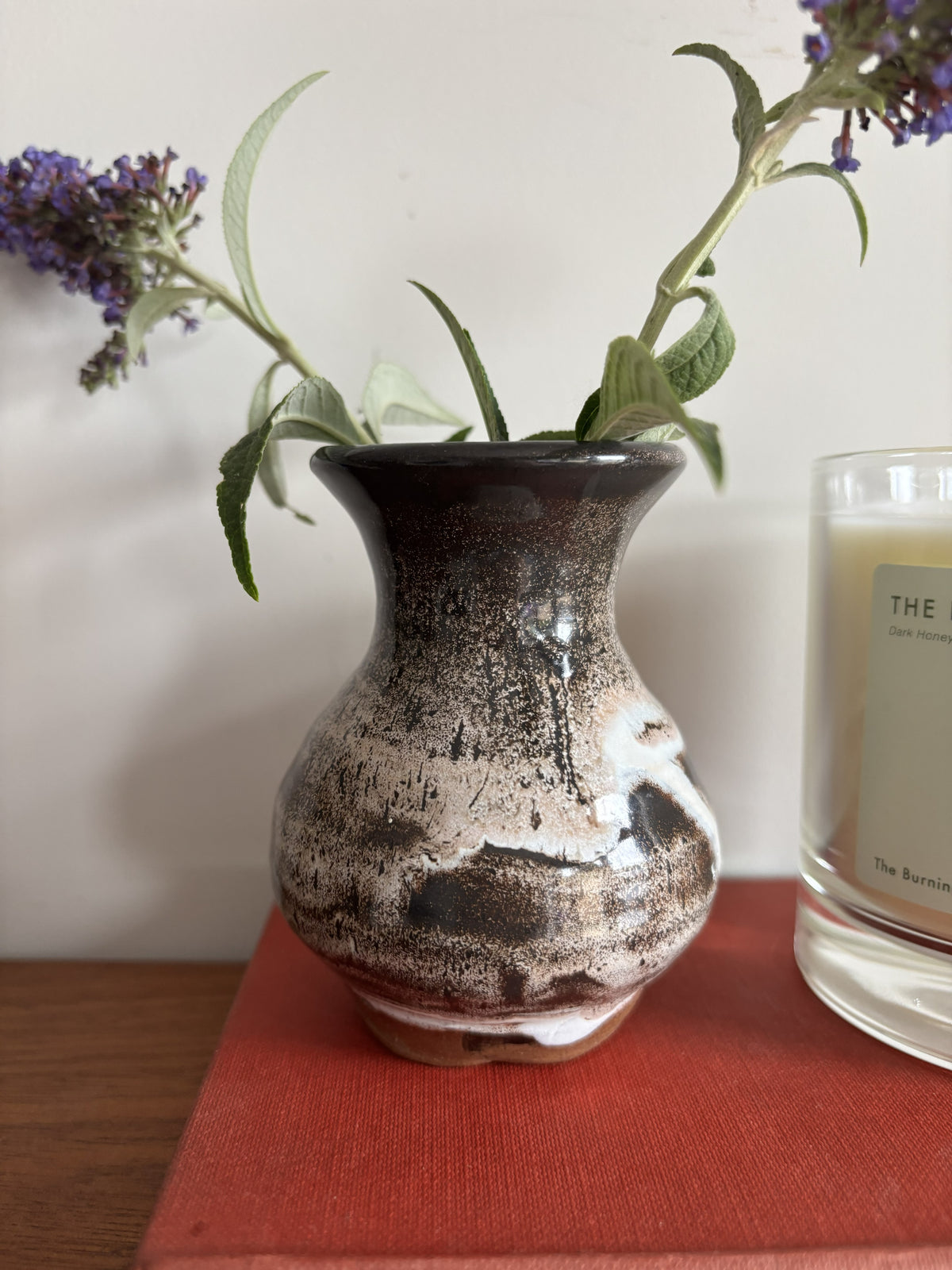 A small handmade ceramic vase with a dark brown, beige, and white glaze pattern, holding green leaves and purple flowers, sitting on a red book next to a candle.