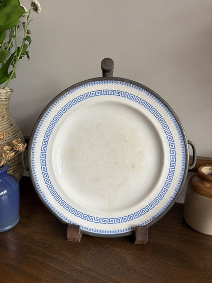 A round white Victorian ceramic plate warmer with a blue Greek key border, displayed on a small stand on a wooden table with decorative plants in the background.