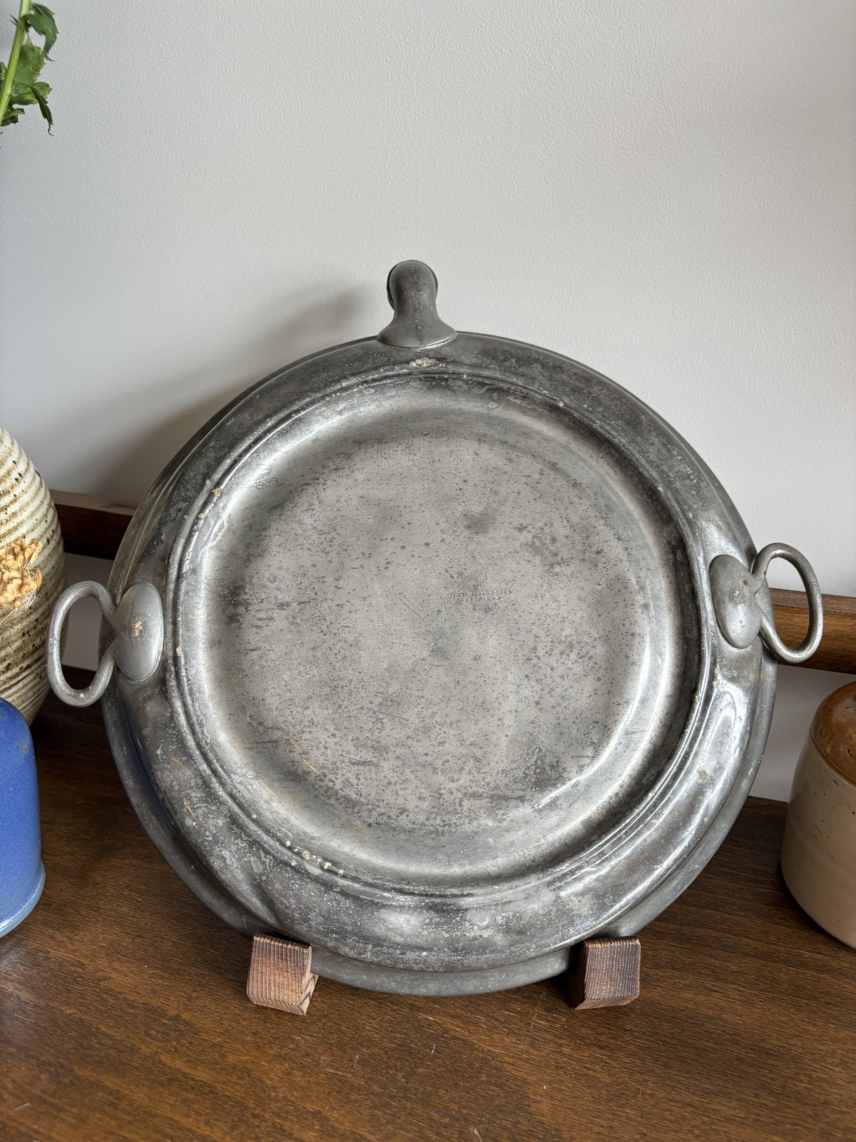 The back of a round Victorian ceramic plate warmer showing the pewter back plate displayed on a small stand on a wooden table with decorative plants in the background