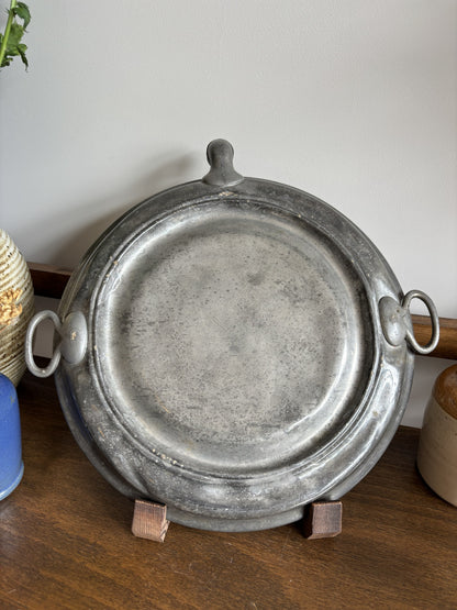 The back of a round Victorian ceramic plate warmer showing the pewter back plate displayed on a small stand on a wooden table with decorative plants in the background