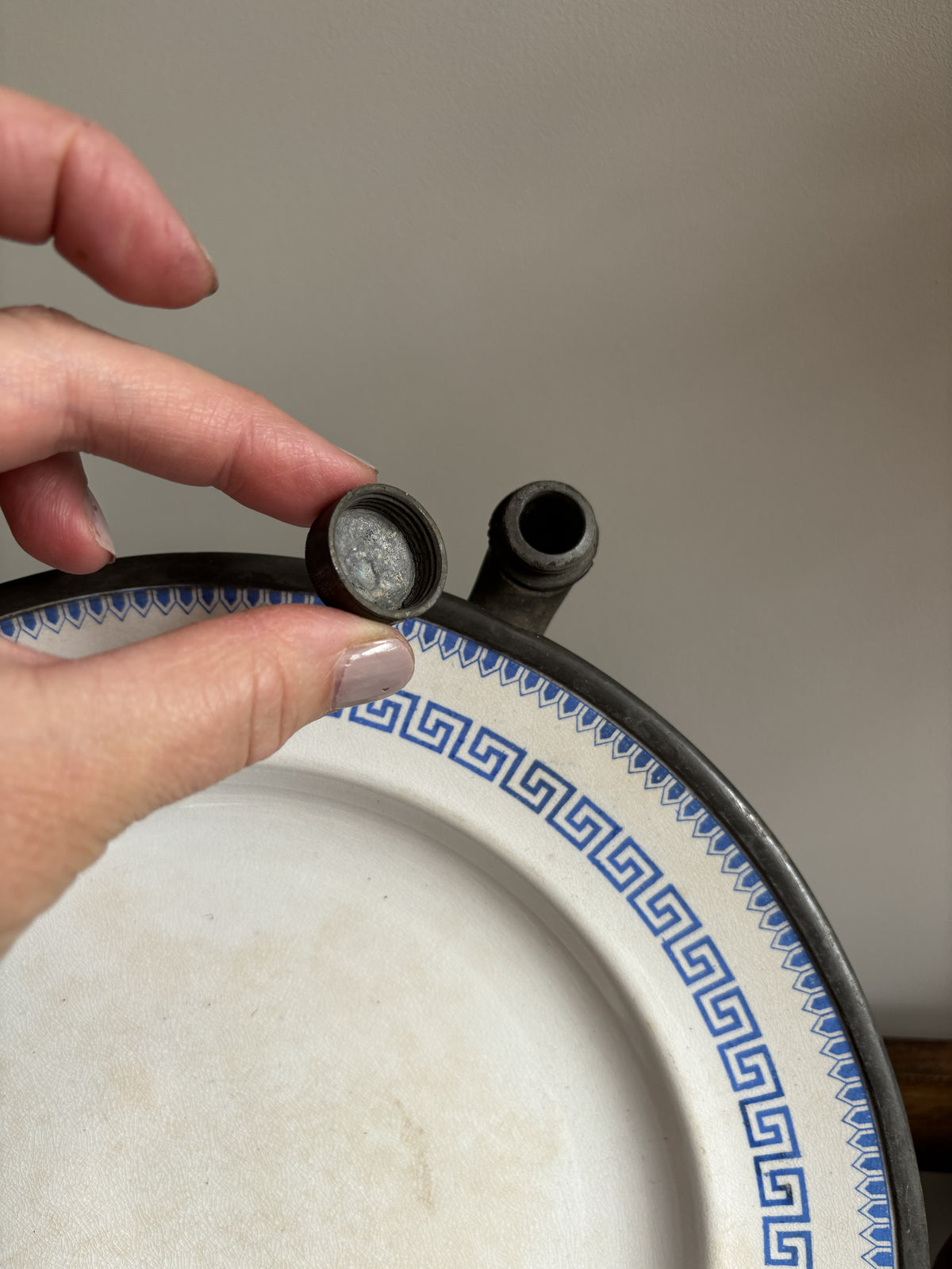 A close-up of a hand holding a small round metal component next to a black cylindrical opening on the rim of a white ceramic plate with a blue Greek key pattern, against a plain beige background.