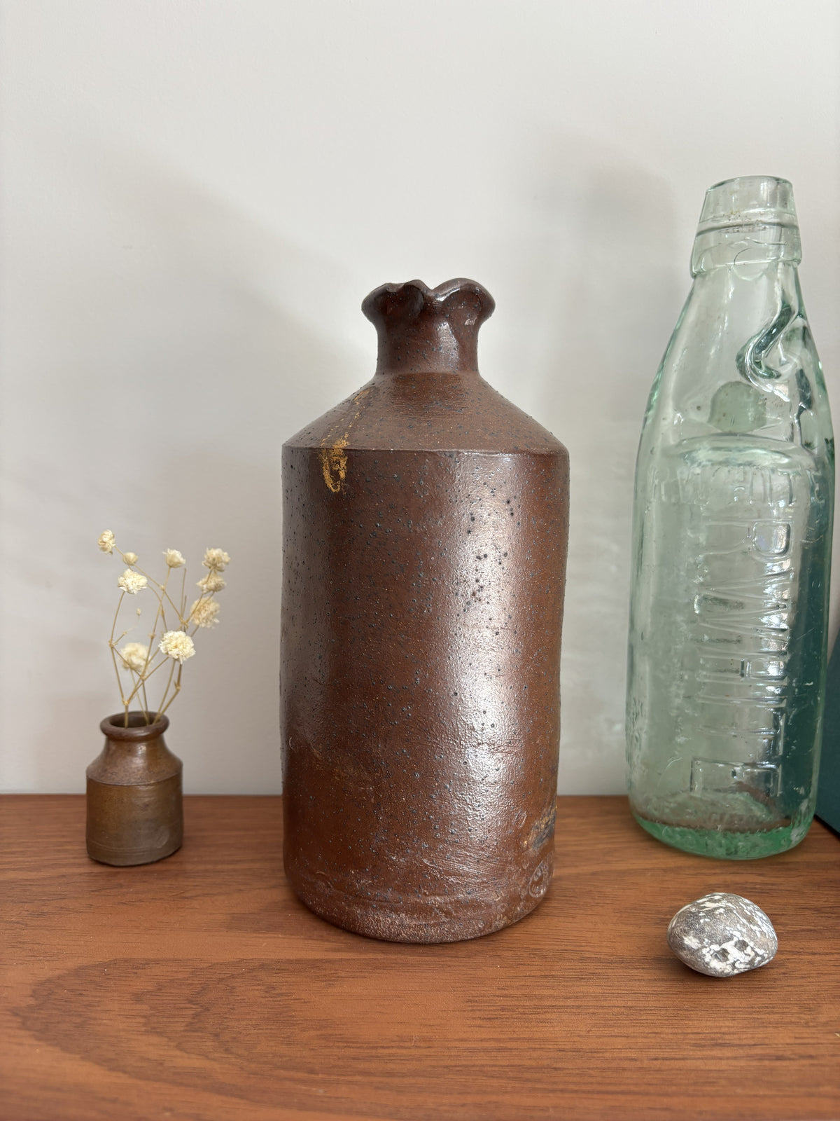 A vintage brown stoneware bottle with a textured, speckled glaze stands on a wooden table against a plain light gray wall. To its left is a small vase with dried white flowers, and to its right is a vintage embossed light green glass bottle with a silver decorative object in front.