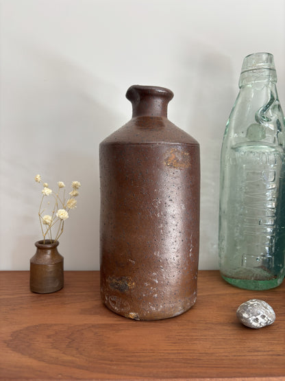 A vintage brown stoneware bottle with a textured, speckled glaze stands on a wooden table against a plain light gray wall. To its left is a small vase with dried white flowers, and to its right is a vintage embossed light green glass bottle with a silver decorative object in front.