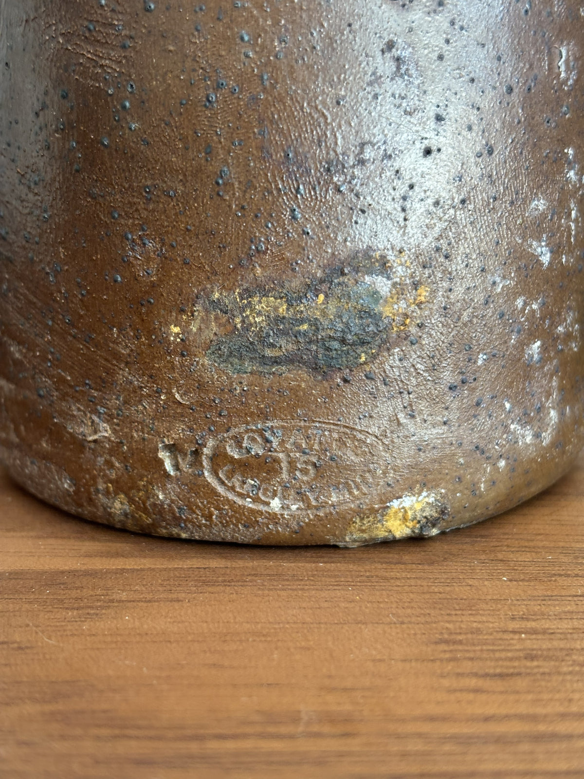 Close-up of the base of a vintage brown stoneware bottle, showing a speckled glaze, a stamped maker's mark, and a small patch of yellow clay repair on a wooden surface.