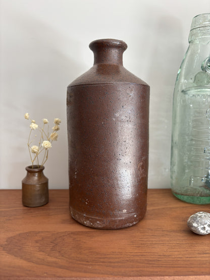 A vintage brown stoneware bottle with a textured, speckled glaze stands on a wooden table against a plain light gray wall. To its left is a small vase with dried white flowers, and to its right is a vintage embossed light green glass bottle with a silver decorative object in front.