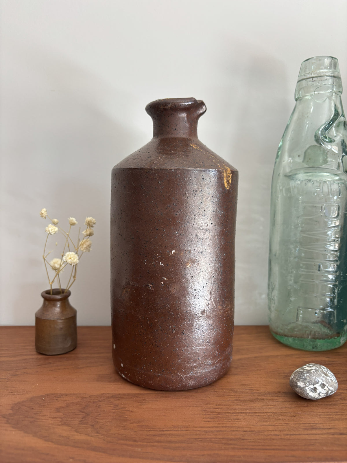A vintage brown stoneware bottle with a textured, speckled glaze stands on a wooden table against a plain light gray wall. To its left is a small vase with dried white flowers, and to its right is a vintage embossed light green glass bottle with a silver decorative object in front.
