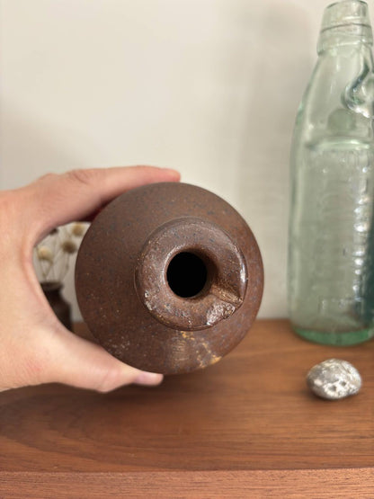 A hand holding a vintage brown stoneware bottle, with a glass bottle and a small silver object on a wooden surface in the background.