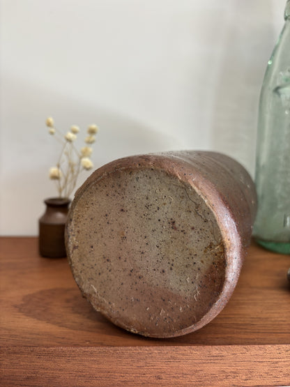 The base of a vintage brown stoneware bottle with a textured, speckled glaze stands on a wooden table against a plain light gray wall. To its left is a small vase with dried white flowers, and to its right is a vintage embossed light green glass bottle with a silver decorative object in front.