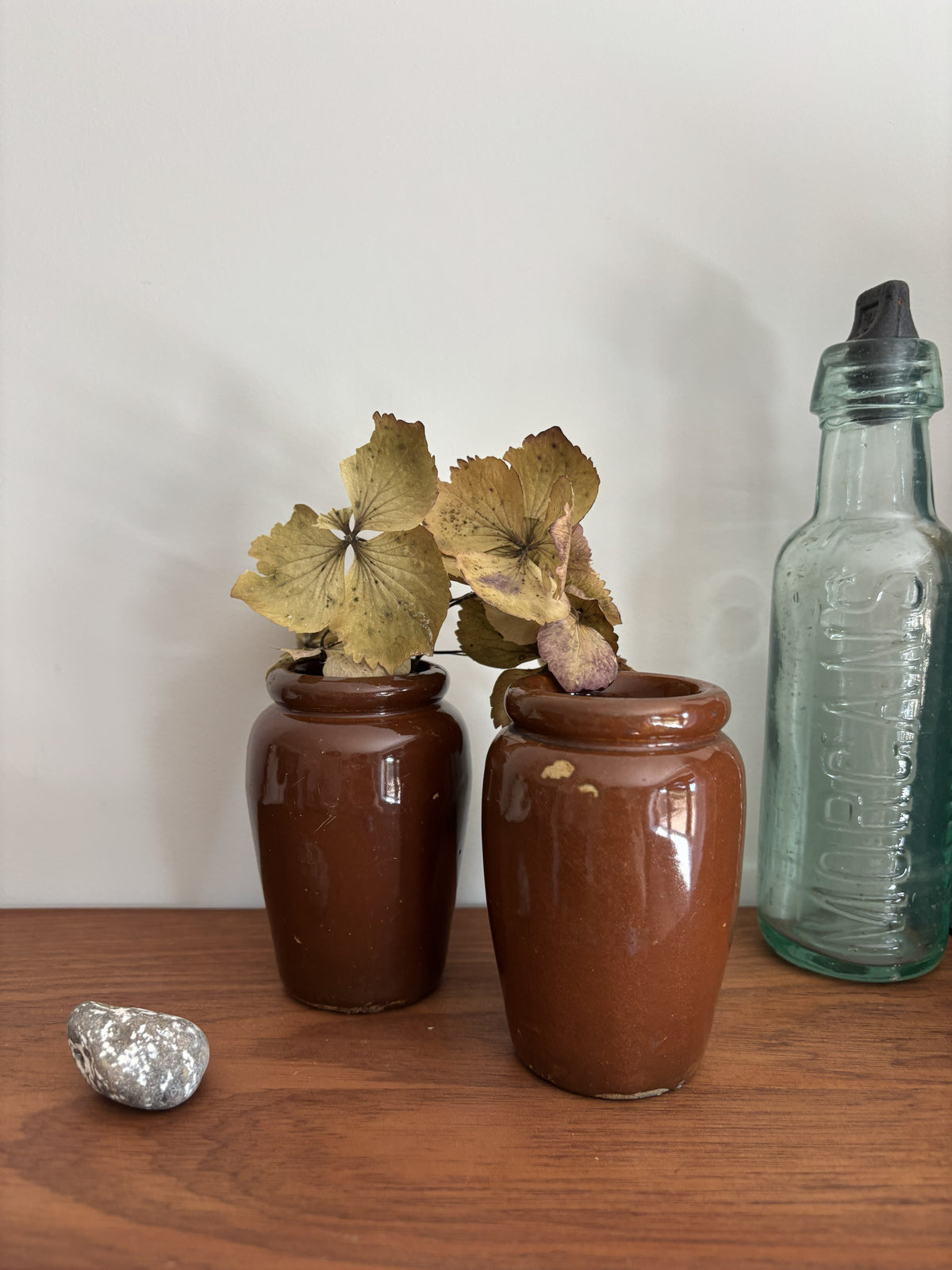 Two small brown ceramic jars with dried flowers growing in them, displayed on a wooden surface next to a vintage glass bottle and a small silver object against a plain wall.