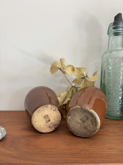 Two vintage brown ceramic bottles lying on their sides on a wooden surface, showing their footprints and glaze marks, with a green glass bottle and dried flowers in the background.
