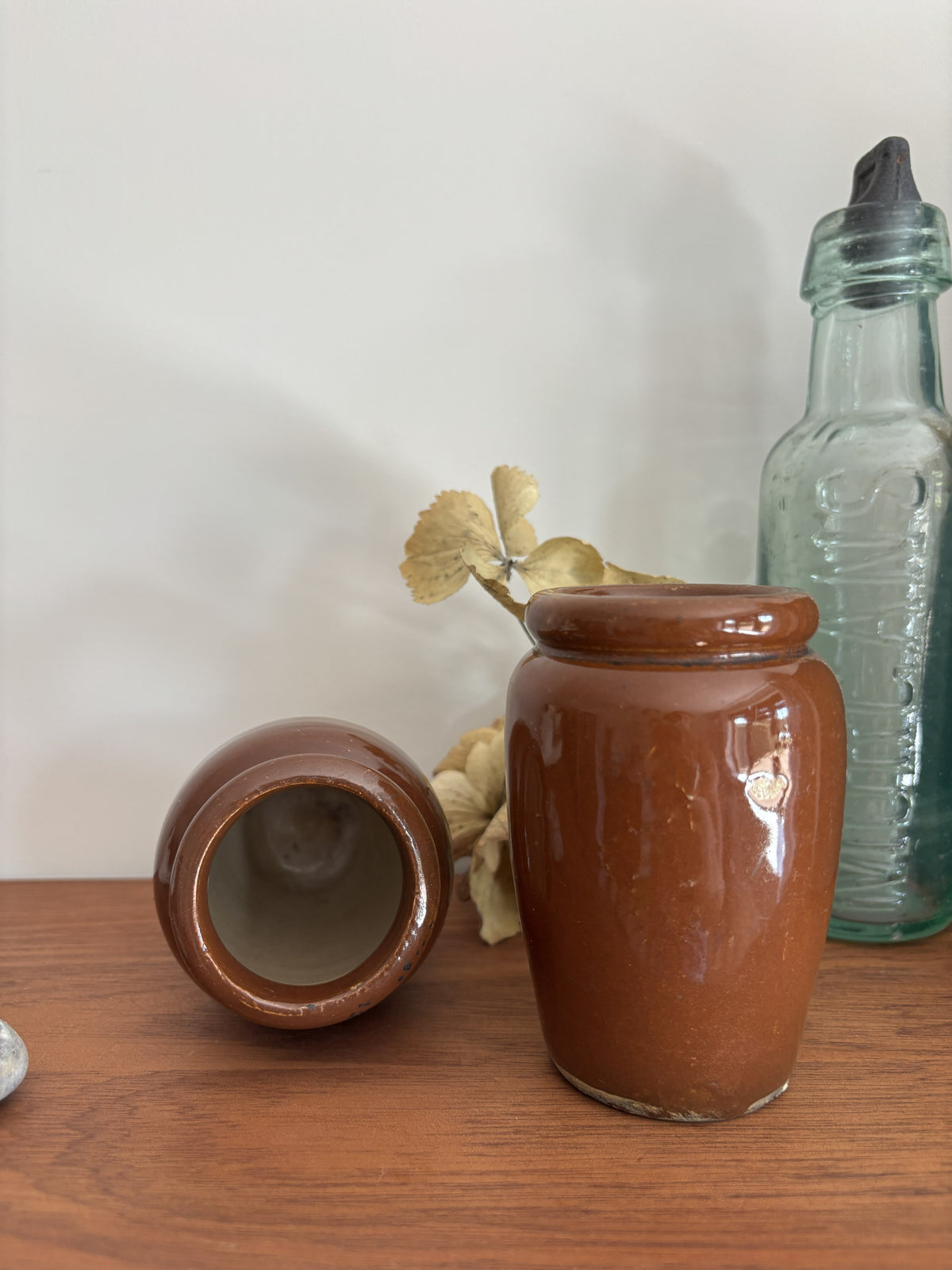 Two small brown stoneware jugs on a wooden surface against a plain light wall, with one jar standing upright and the other lying on its side to show the inside. A translucent green glass bottle and dried leaves sit behind them as props.