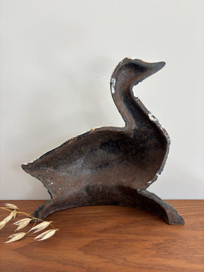 The reverse of a vintage cast iron duck doorstop, displayed on a wooden tabletop against a plain light gray wall with dried wheat stalks in the foreground.