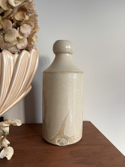 A vintage stone bottle with a rounded shoulder and a textured cream-beige glaze, displayed on a wooden surface next to a smaller stoneware jar and dried flowers.