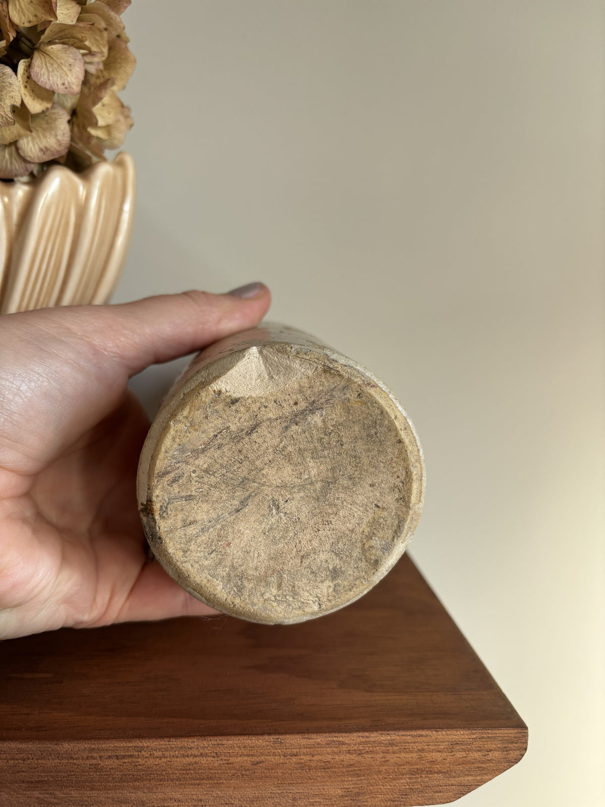 A hand holding a vintage cream stoneware bottle showing a textured, weathered surface and a chip. The background features a neutral beige wall and a wooden surface.