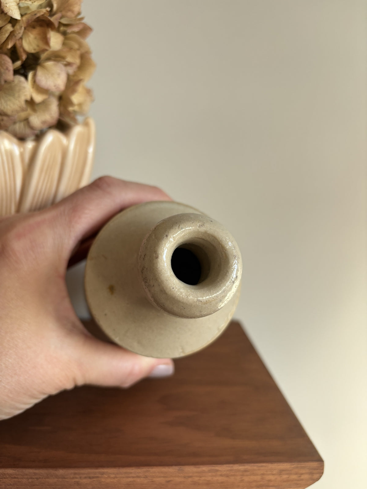 A hand holding the top opening of a cream vintage stoneware bottle, with dried flowers in a beige pot and a wooden surface in the background.