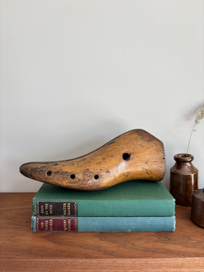 A vintage wooden shoe last with circular holes rests on top of two old books on a wooden table, with a small ceramic vase in the background against a light gray wall.