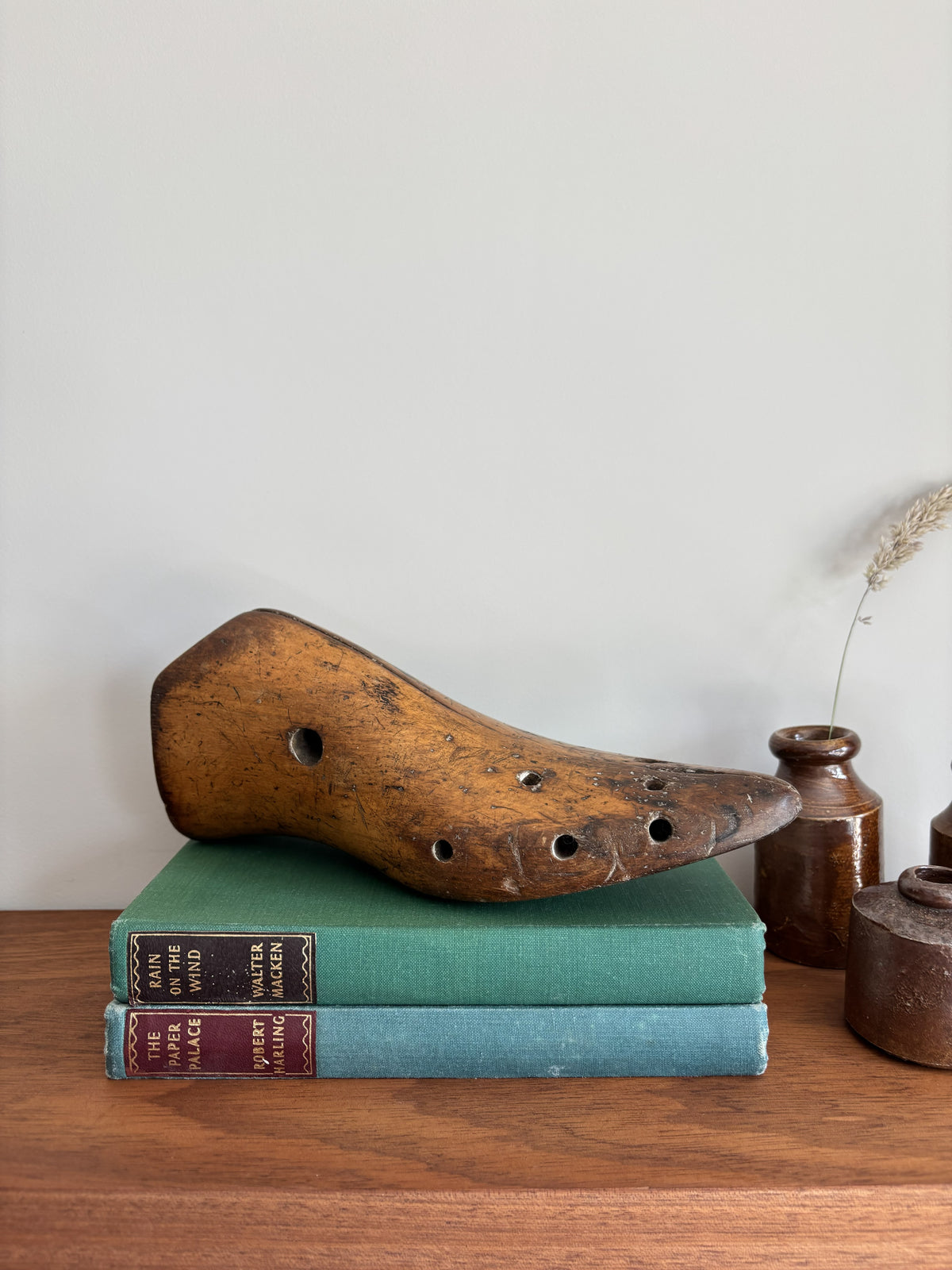 A rustic wooden shoe last with multiple holes rests on top of two vintage hardcover books on a wooden surface, with small ceramic vases in the background against a plain wall.