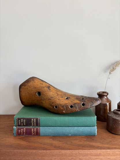 A rustic wooden shoe last with multiple holes rests on top of two vintage hardcover books on a wooden surface, with small ceramic vases in the background against a plain wall.