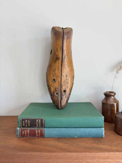 A vintage wooden shoe last stands upright on top of two stacked antique books on a wooden table against a plain light wall.