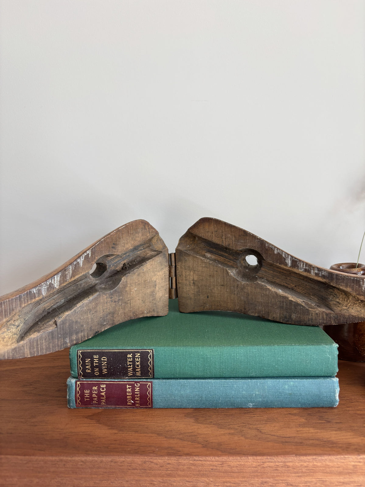 A vintage wooden shoe last with metal pivot pins, displayed on top of two vintage hardcover books on a wooden surface against a plain light gray wall.