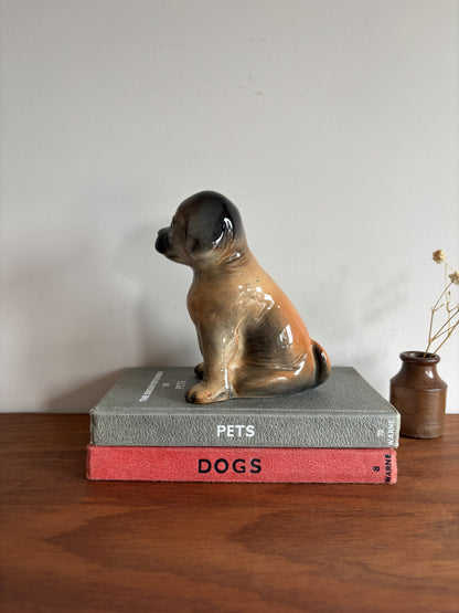 A glossy ceramic puppy dog figurine in tan, brown, and black sits on top of two stacked books on a wooden surface against a plain wall.