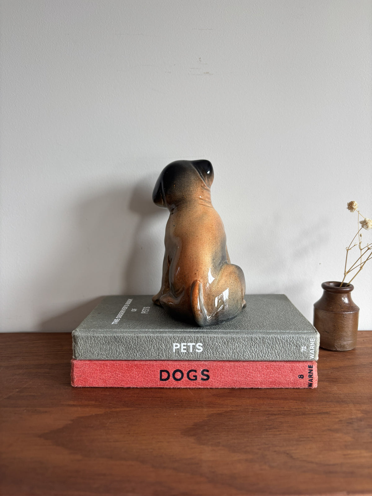 A glossy ceramic puppy dog figurine in a sitting position, displayed on top of two stacked books on a wooden surface against a plain wall.