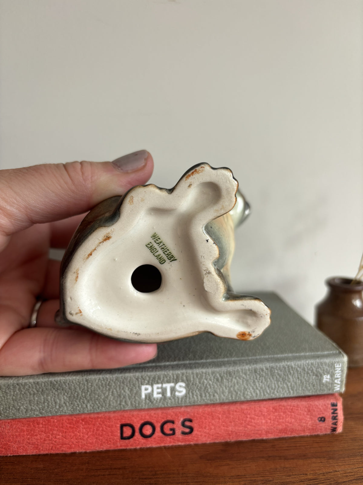 A hand holds a small ceramic dog figurine upside down on top of two books, one gray with the word 'PETS' and one red with the word 'DOGS', against a plain indoor background.  The words Wetherby England are stamped in green on the base. 