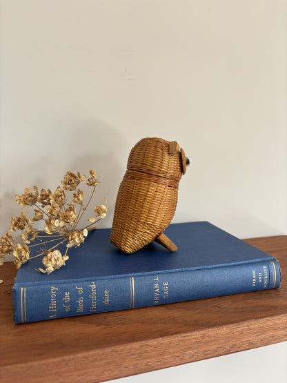 A small woven owl-shaped basket in natural wicker sits on top of a blue hardcover book, with dried flowers beside it on a wooden surface against a plain light wall.