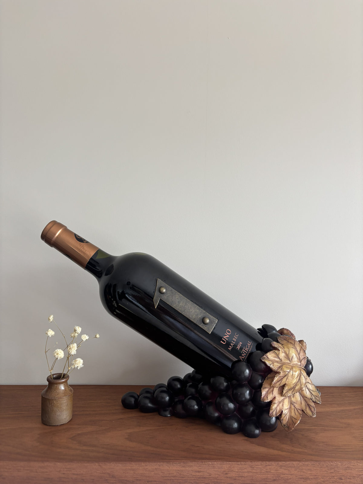 A decorative wine bottle sculpture lying on its side, surrounded by dark grapes and a metallic gold grapevine leaf, displayed on a wooden surface next to a small vase with dried white flowers against a plain light gray wall.