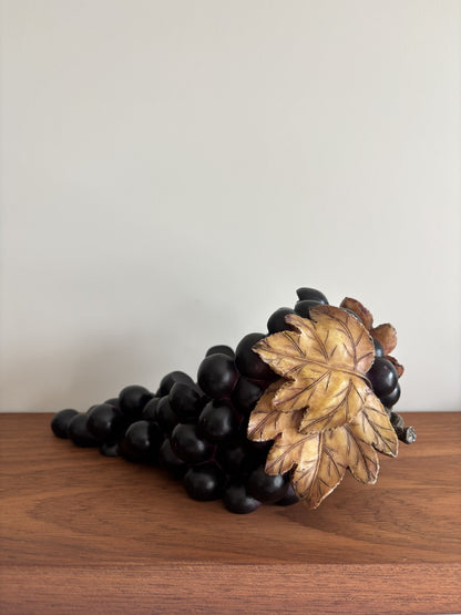 A decorative sculpture of a grapevine with dark purple-black grapes and a single gold-toned leaf, displayed on a wooden surface against a plain light gray wall.