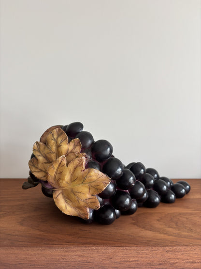 A decorative sculpture of a grapevine with dark purple-black grapes and two golden-brown leaves, displayed on a wooden surface against a plain light gray wall.