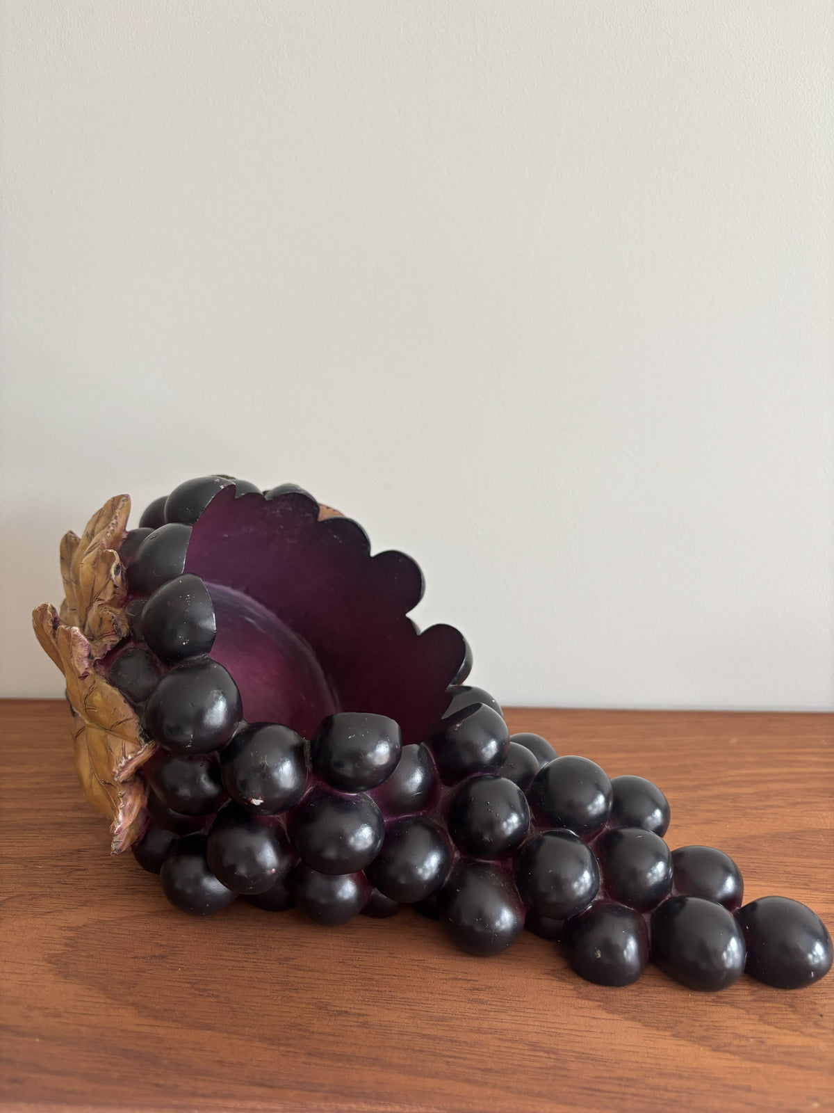 A decorative sculpture of a grapevine or cornucopia-like form made of glossy dark purple and black grape-like spheres, with a brown textured base, displayed on a wooden surface against a plain light wall.