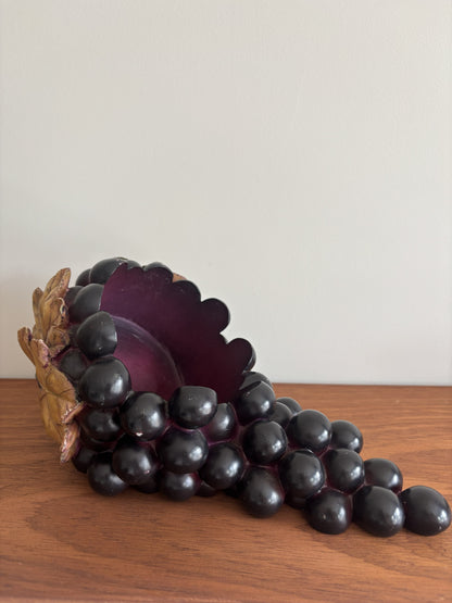 A decorative sculpture of a grapevine or cornucopia-like form made of glossy dark purple and black grape-like spheres, with a brown textured base, displayed on a wooden surface against a plain light wall.