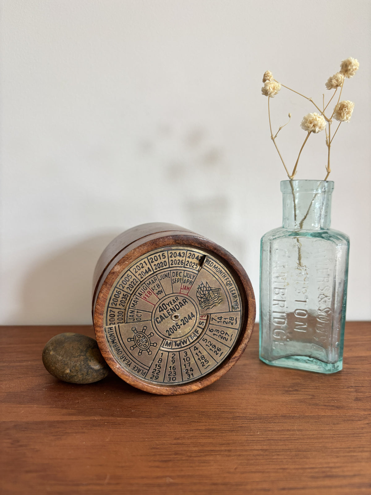 A vintage wooden calendar box with a detailed calendar dial, lying on its side on a wooden table next to a small stone and a light blue glass bottle with dried flowers.