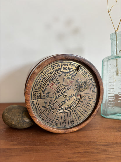 A round wooden 40-year calendar stand displaying the calendar details for 2006-2046, styled on a wooden tabletop with a stone and a vintage glass bottle in the background.