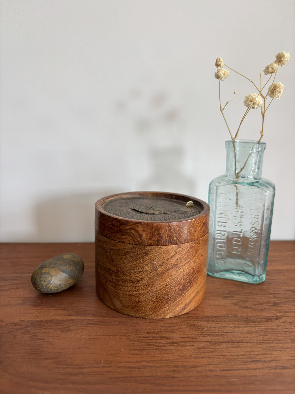 A round wooden box with a lid sits on a wooden table next to a small stone and a vintage-style light blue glass bottle holding dried cream flowers, against a plain light wall.