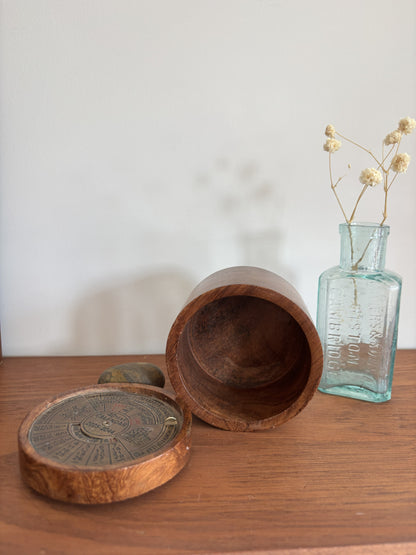 A rustic wooden box with its lid placed beside it on a wooden table, next to a small vintage glass bottle holding dried flowers against a plain light wall.