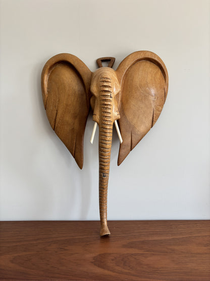 A carved wooden elephant wall hanging with large ears, a textured trunk, and small wooden tusks, displayed against a plain light wall above a wooden surface.
