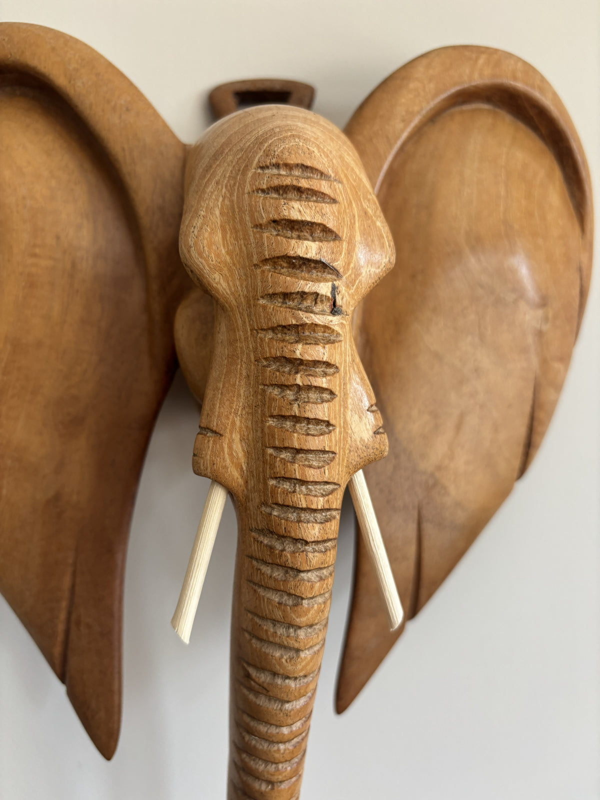 A carved wooden elephant wall decor sculpture with large ears, a textured trunk, and small ivory-colored tusks, shown against a plain light background.
