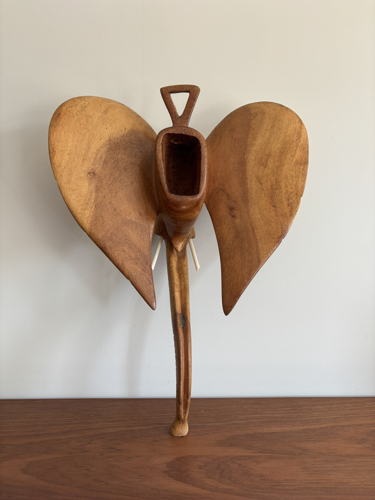 A handcrafted wooden sculpture of an elephant with large wings, displayed on a wooden surface against a plain off-white wall.