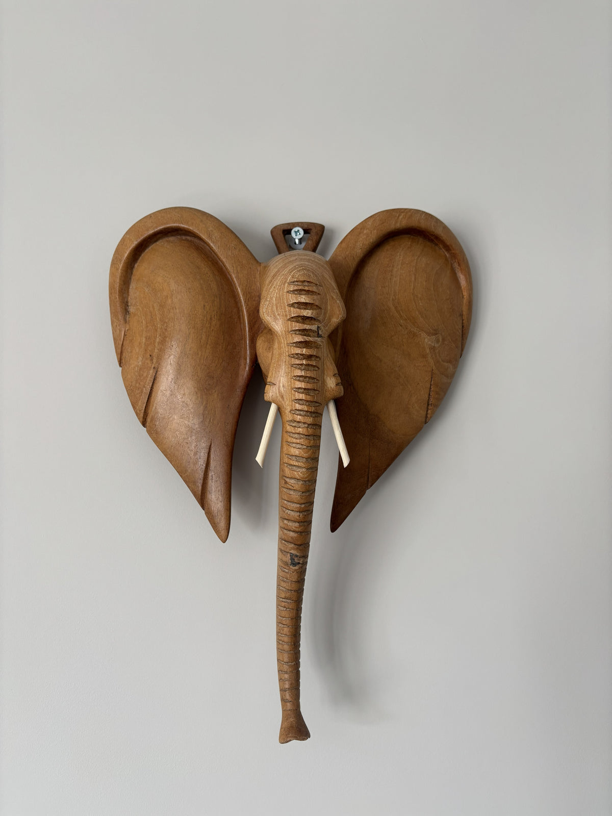 A carved wooden elephant wall sculpture with large ears, a trunk, and small tusks, mounted against a plain light gray wall.