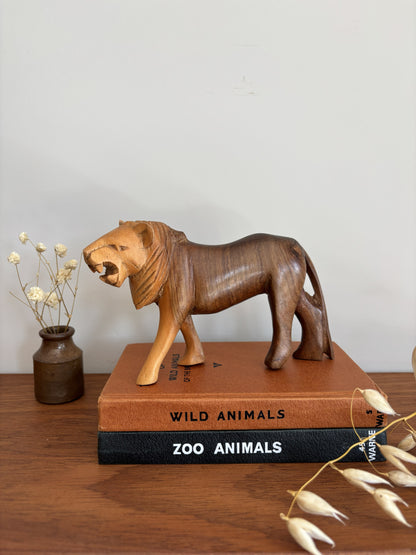 A carved wooden lion figurine with a roaring expression, displayed on top of two books on a wooden surface. The books are labeled 'WILD ANIMALS' and 'ZOO ANIMALS'.