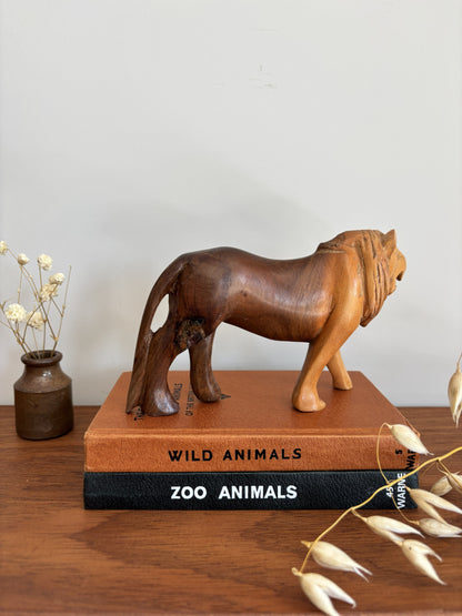 A carved wooden lion figurine in a medium brown finish stands on top of two stacked books, one tan and one black, on a wooden tabletop. A small vase with dried stems sits to the left, and dried plant pods hang in the foreground against a plain light wall.